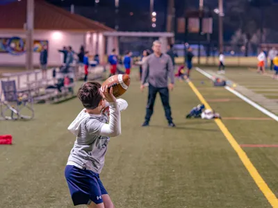 Young boy winding up to throw a football on a lit turf field during an evening practice