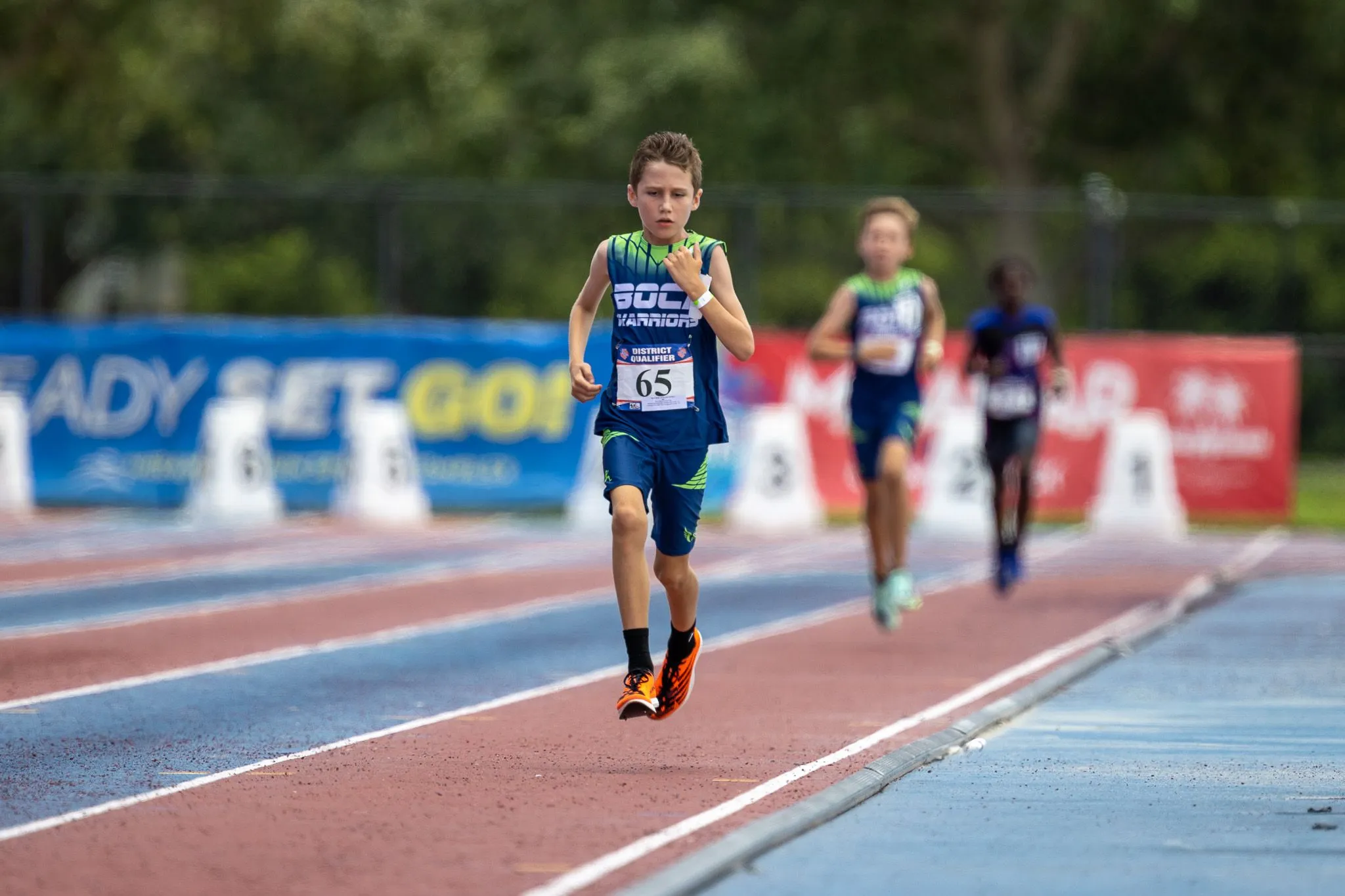 Boy in Boca Warriors jersey crossing the finish area ahead of competitors at a track meet