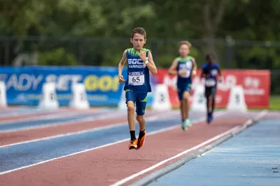 Boy in Boca Warriors jersey crossing the finish area ahead of competitors at a track meet