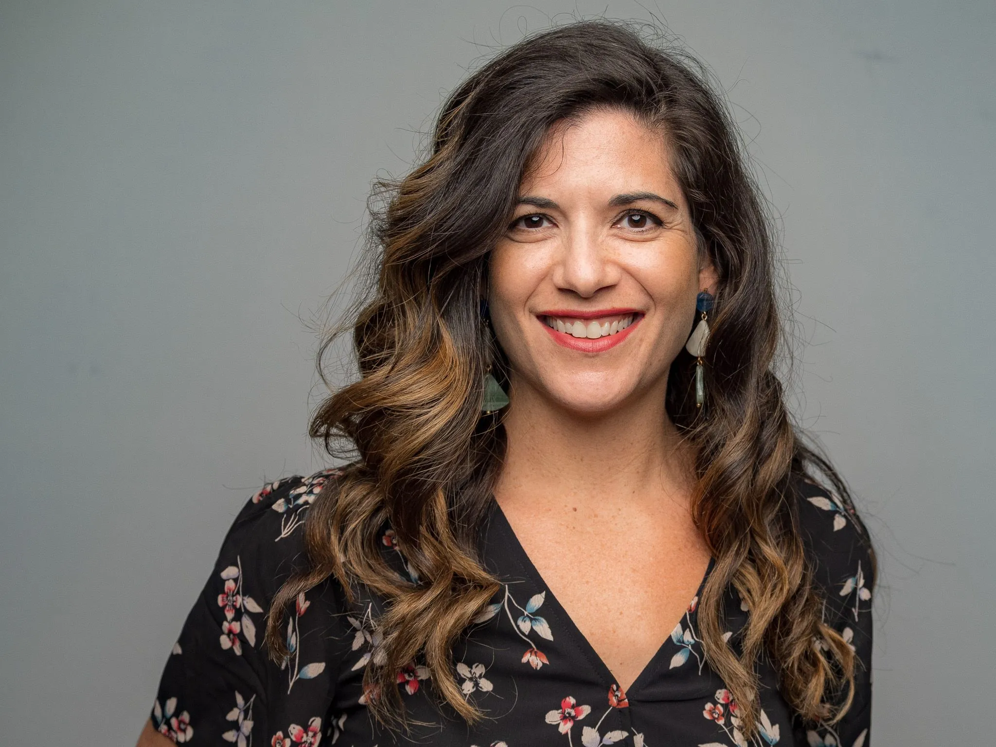 Woman with curly brown hair and statement earrings smiling in a floral blouse on gray backdrop