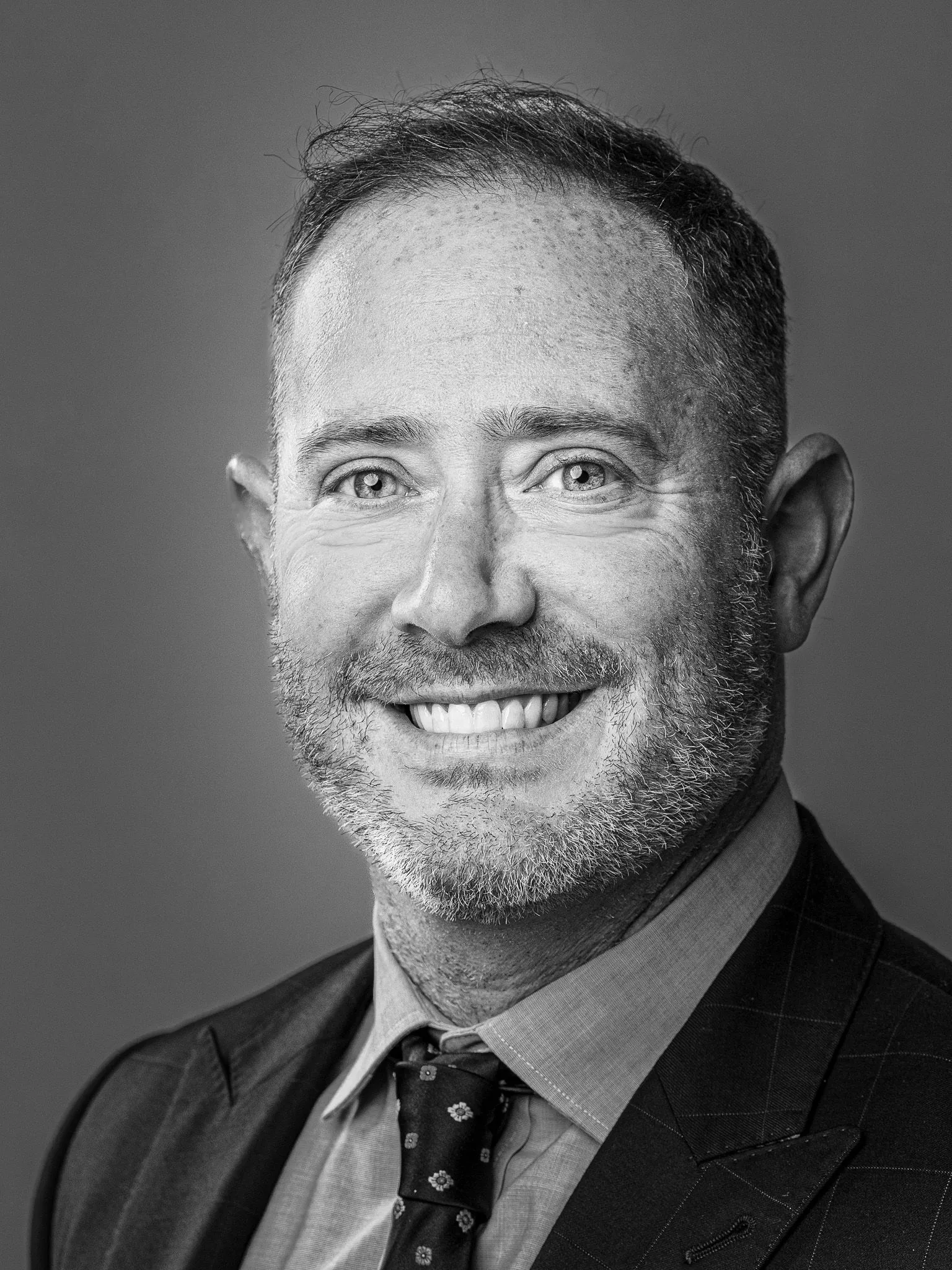 Black and white headshot of a man in a plaid suit and patterned tie with a warm smile