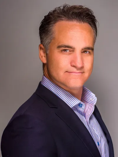 Man in a navy blazer and striped shirt posing for a professional headshot on gray background