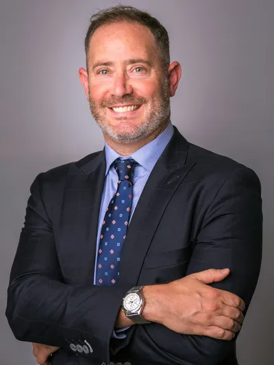 Man in a navy suit with arms crossed smiling confidently against a gray studio backdrop