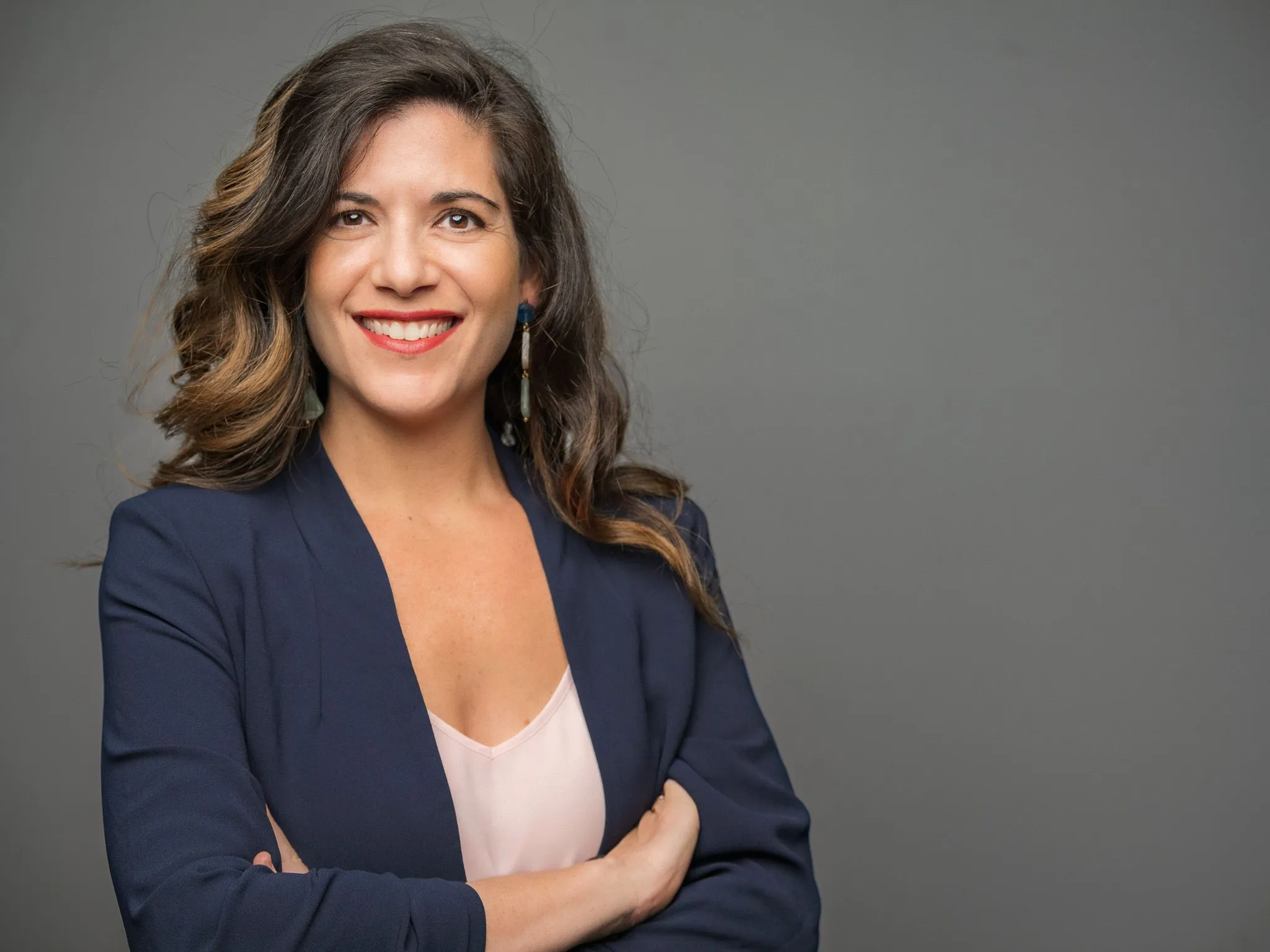 Woman with wavy brown hair in a navy blazer smiling with arms crossed on gray background