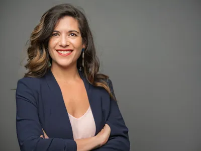 Woman with wavy brown hair in a navy blazer smiling with arms crossed on gray background