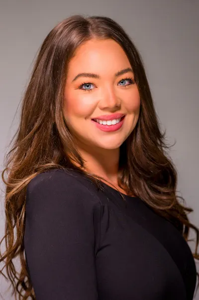 Young woman with long brown hair smiling in a black top against a gray studio backdrop