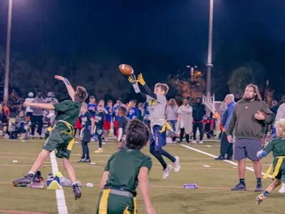 Youth flag football player leaping to catch a pass under the lights with crowd watching