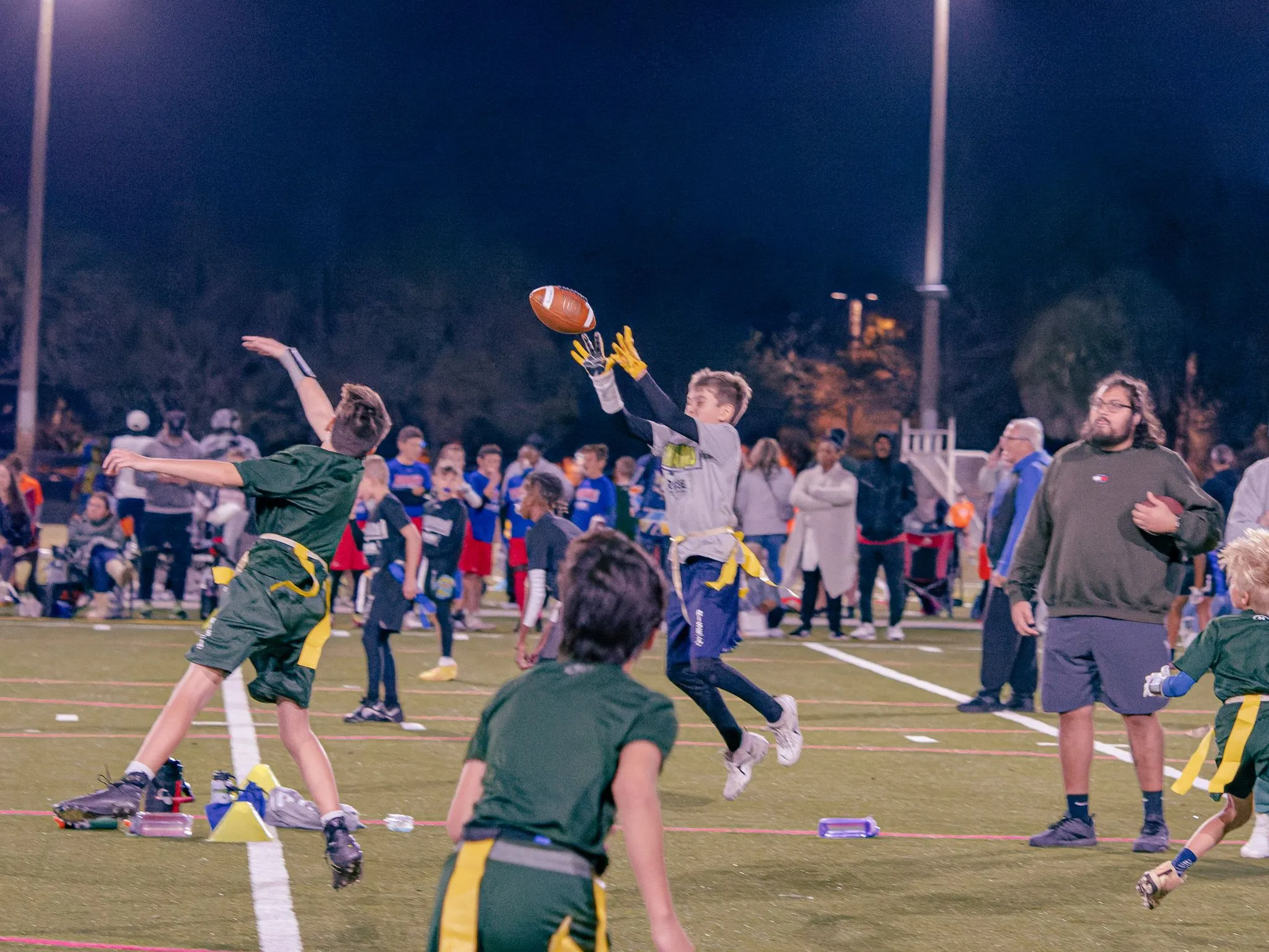 Youth flag football player leaping to catch a pass under the lights with crowd watching
