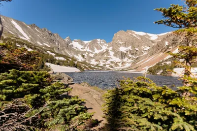 Snow-patched mountain cirque framed by evergreen trees with an alpine lake in the foreground