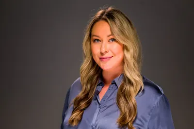 Blonde woman in a satin blue blouse posing for a professional headshot on gray backdrop