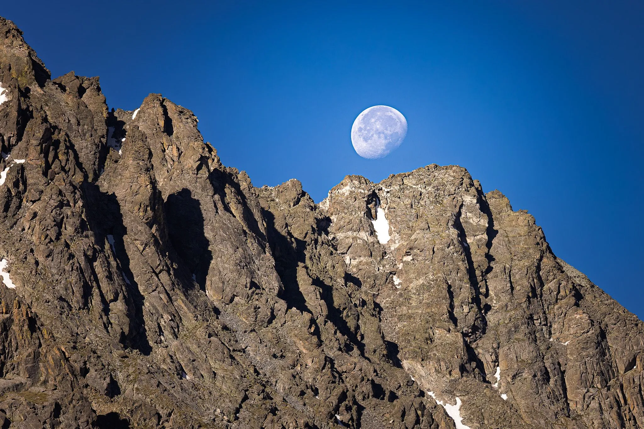 Nearly full moon rising over a jagged mountain ridgeline against a deep blue sky