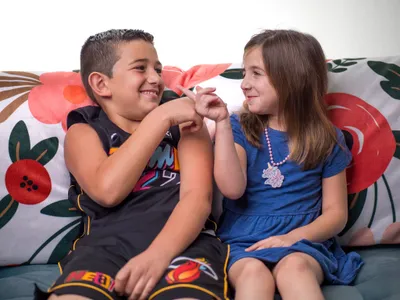 Brother and sister sitting on a couch smiling at each other with a playful finger gesture