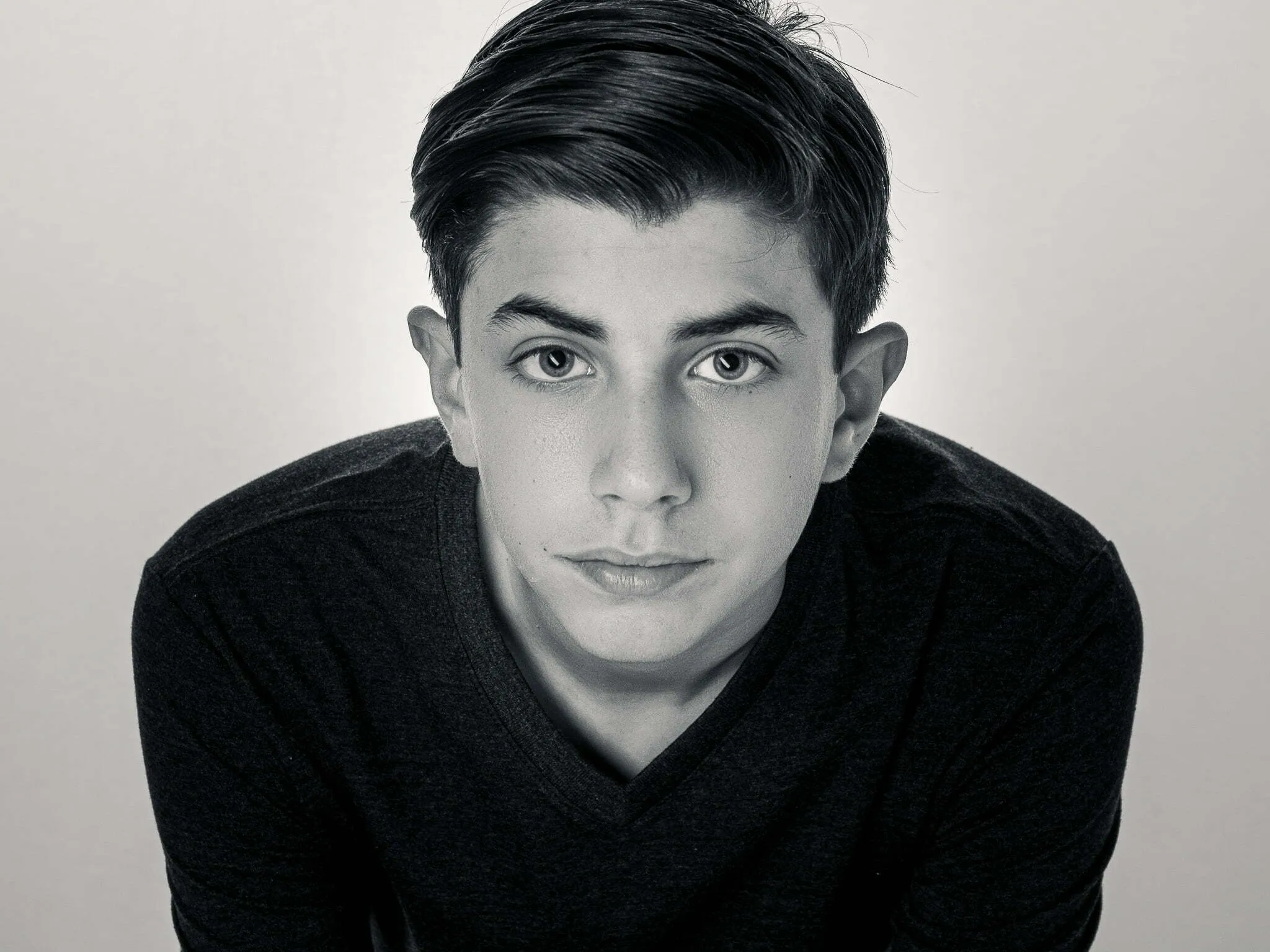 Teen boy in black v-neck shirt looking directly at camera, black and white studio headshot