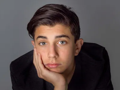 Teen boy resting chin on hand in dark jacket, color studio headshot with gray background
