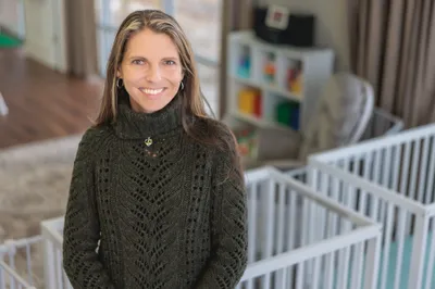 Woman in her 40s wearing dark green knit sweater smiling warmly in a bright childcare space with cribs