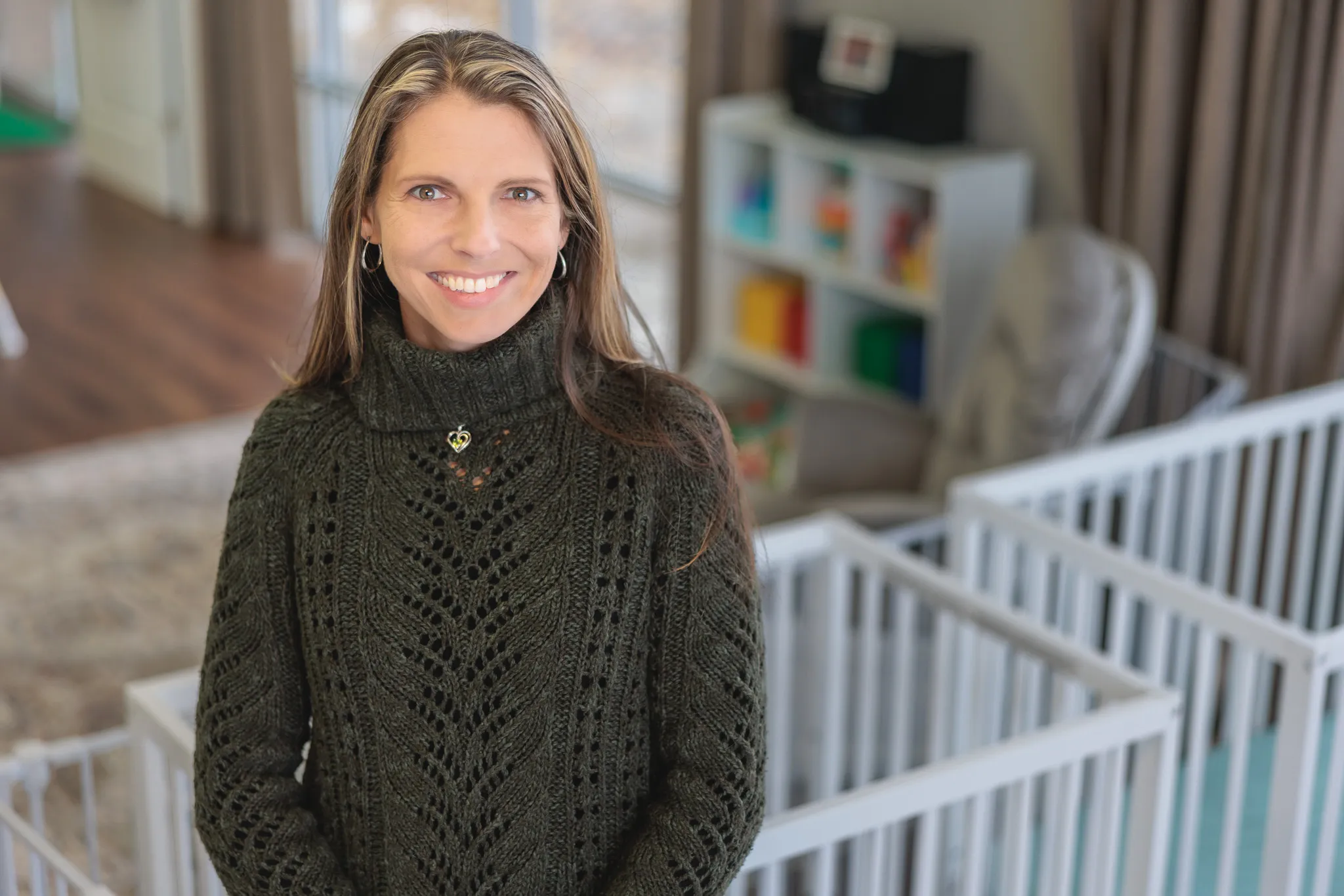 Woman in her 40s wearing dark green knit sweater smiling warmly in a bright childcare space with cribs