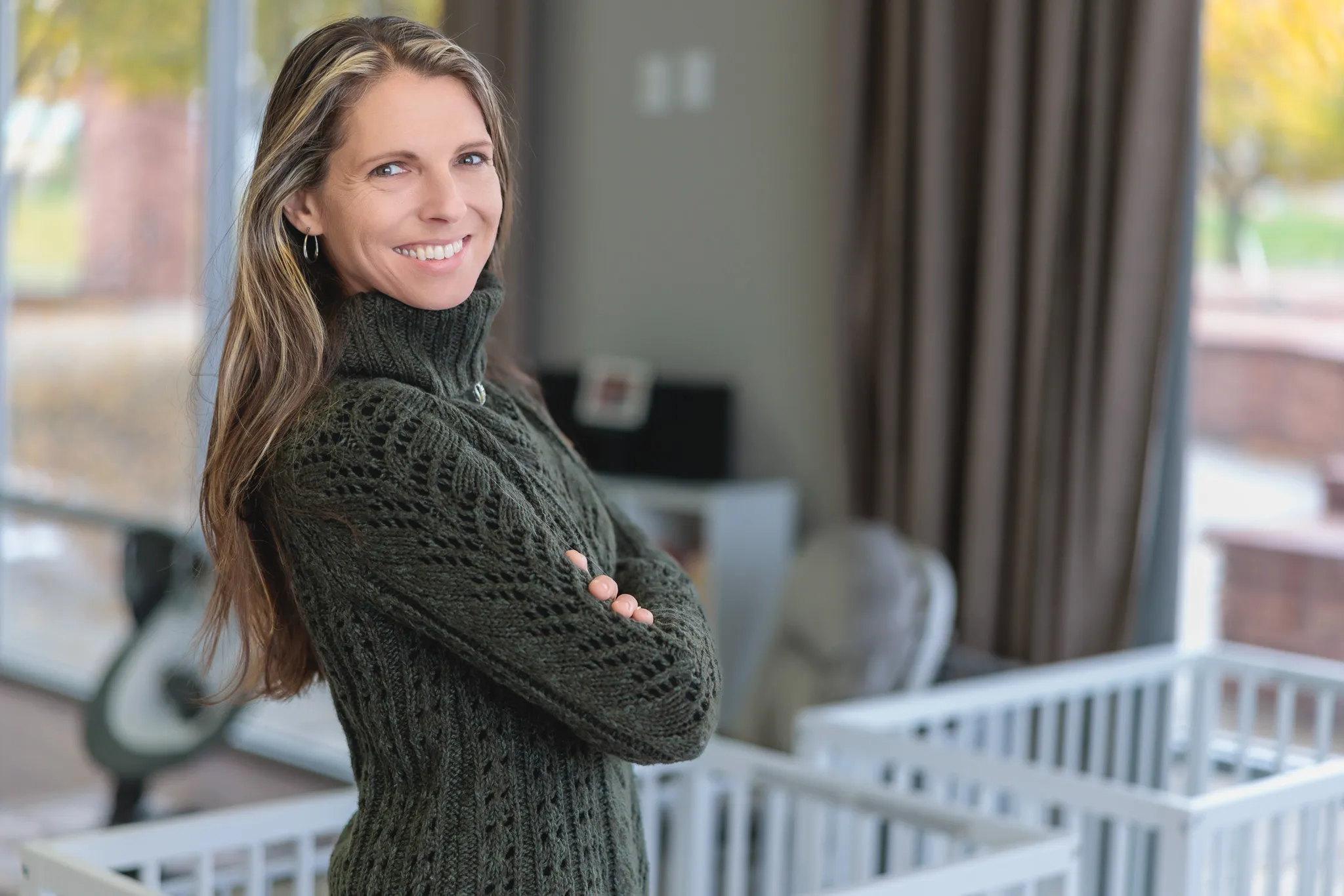 Woman with arms crossed smiling confidently on a covered porch with autumn trees visible in the background