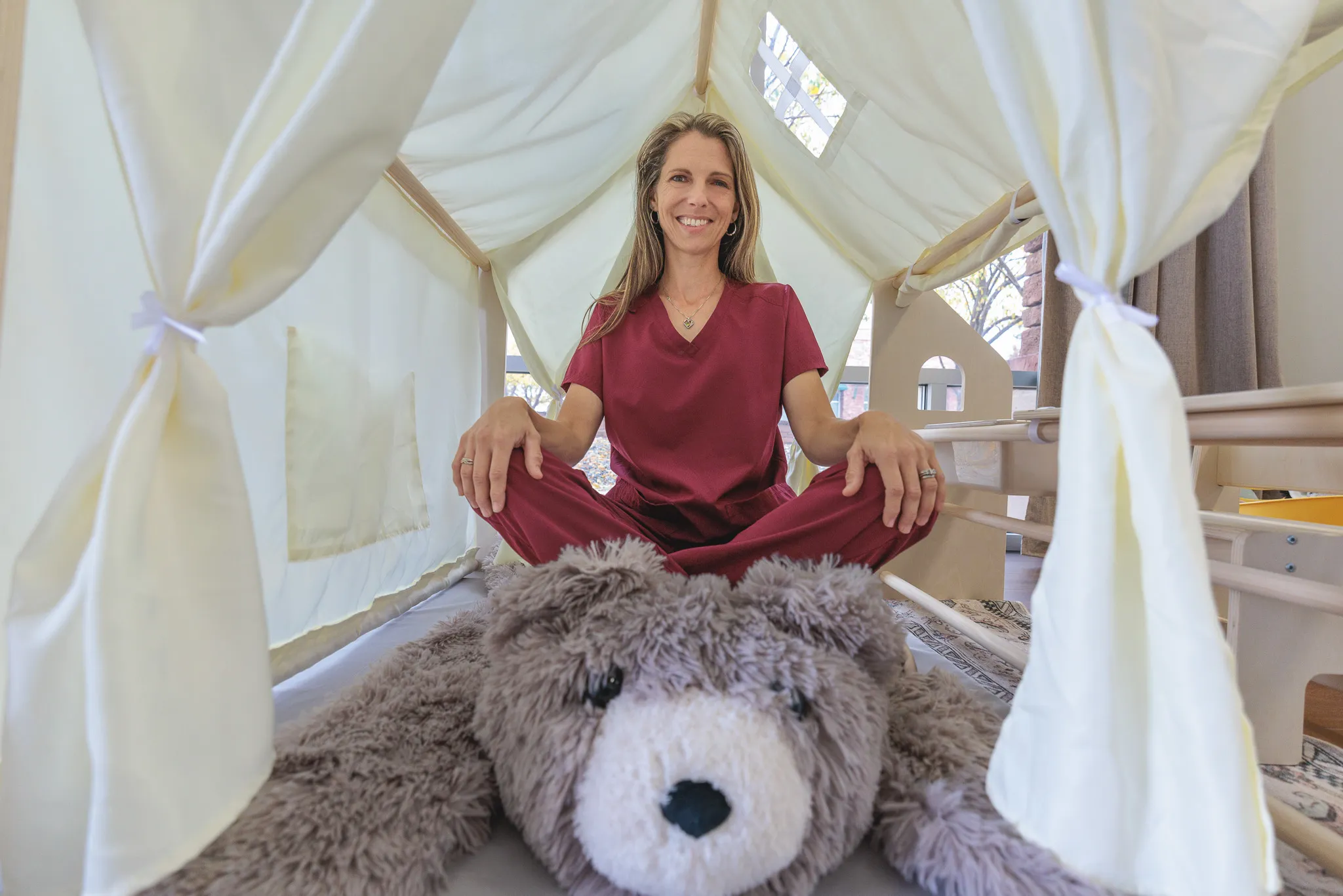 Woman in burgundy scrubs sitting cross-legged inside a white canopy tent with a large stuffed bear in foreground