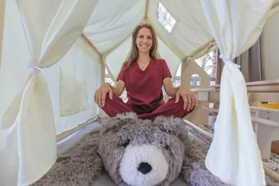 Woman in burgundy scrubs sitting cross-legged inside a white canopy tent with a large stuffed bear in foreground