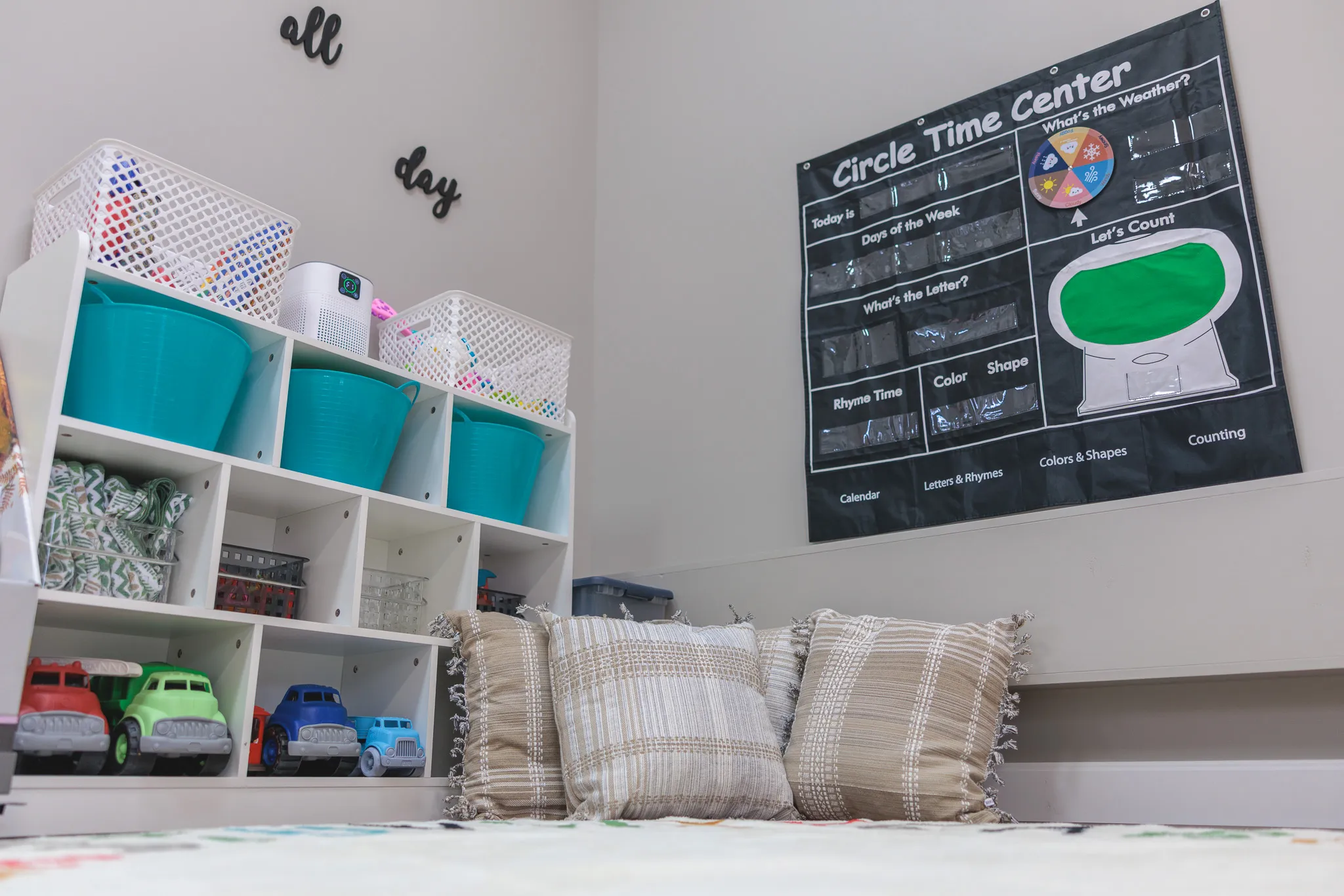 Daycare reading nook with cubby shelves, teal bins, toy trucks, plaid pillows, and Circle Time Center poster