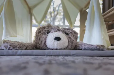 Close-up of a fluffy brown teddy bear lying flat inside a white fabric play tent with soft natural light