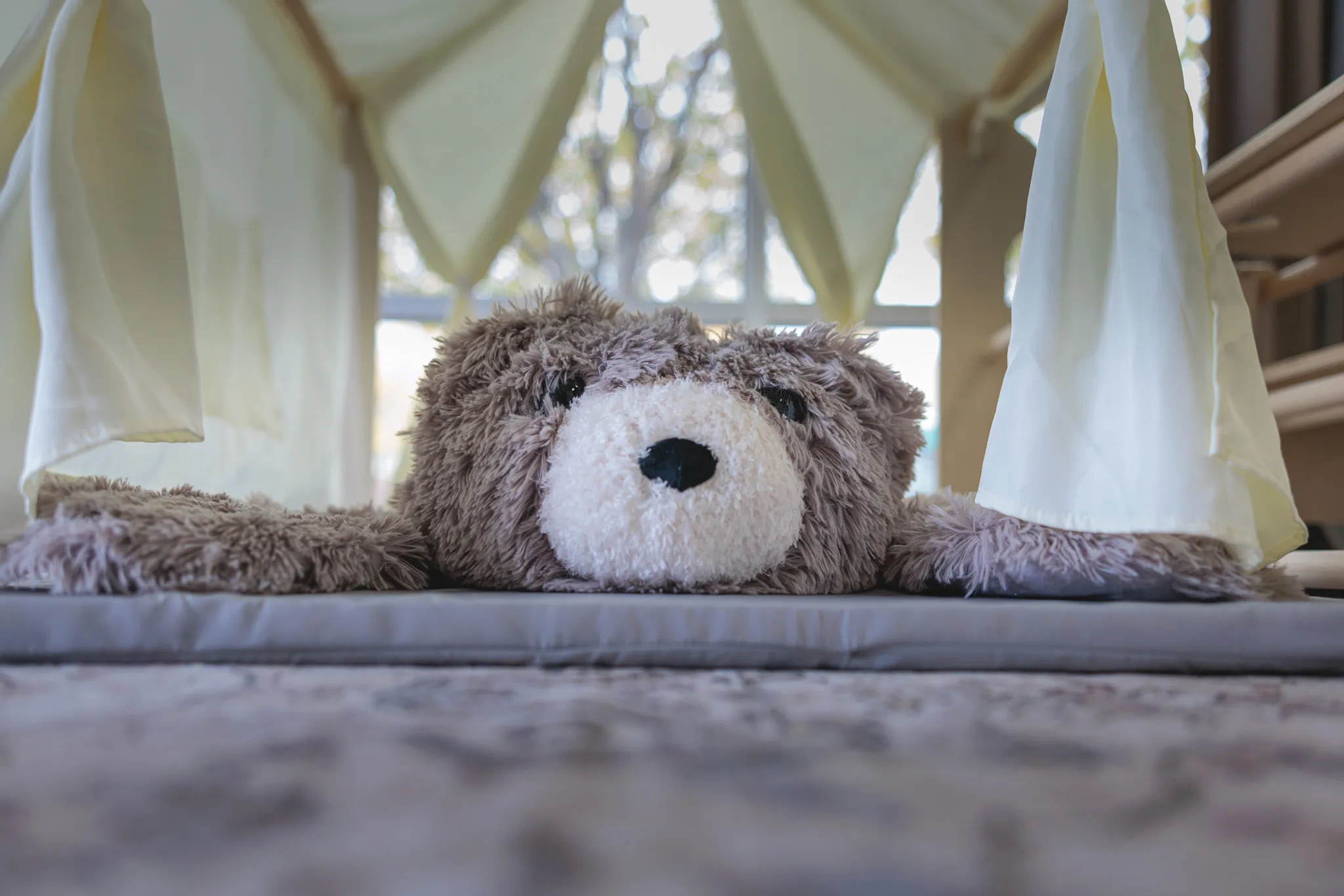 Close-up of a fluffy brown teddy bear lying flat inside a white fabric play tent with soft natural light