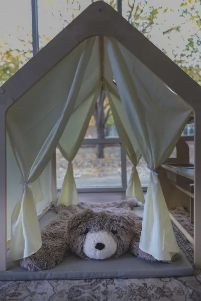 Wooden house-shaped play tent with white curtains and stuffed bear inside, autumn trees through window behind