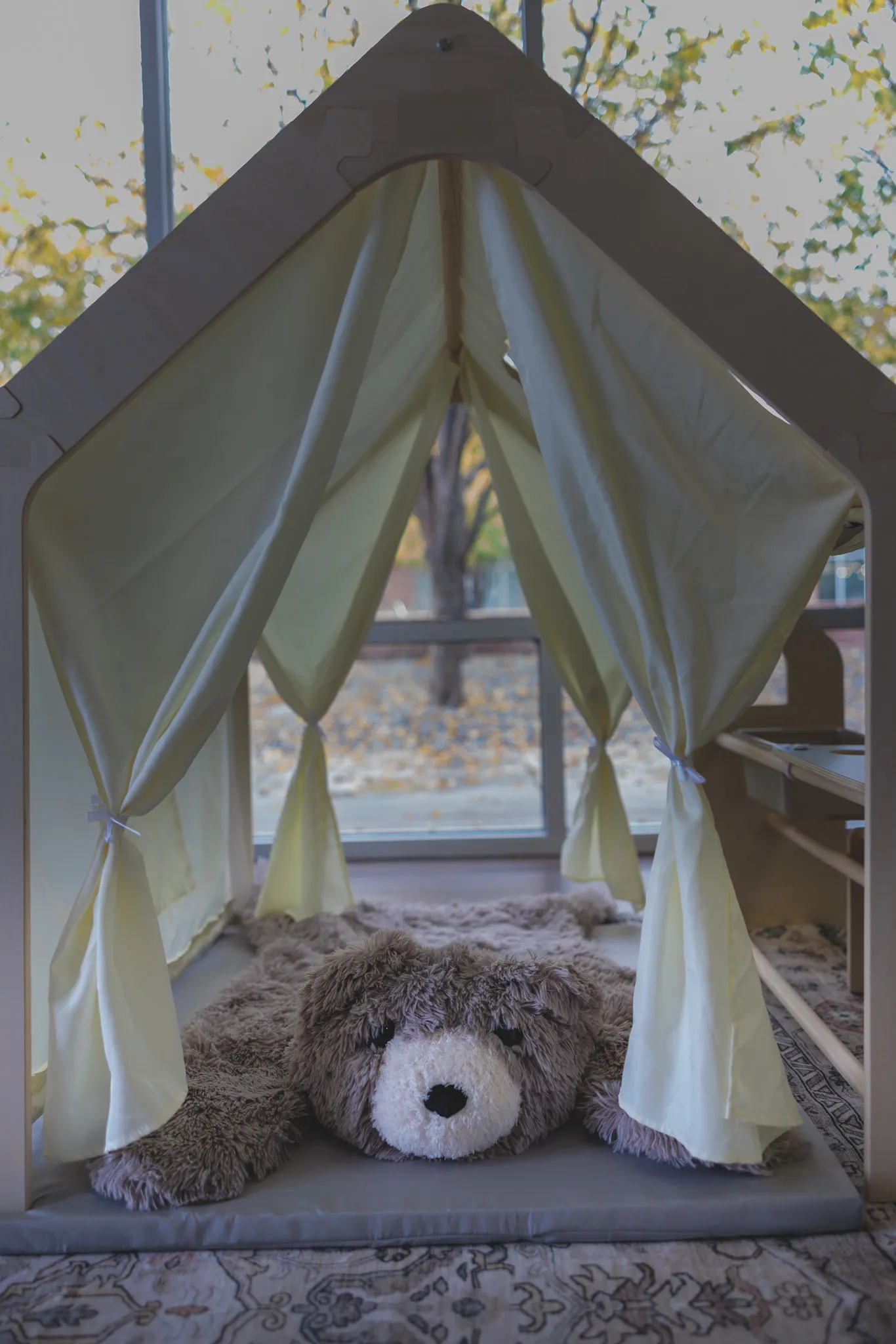 Wooden house-shaped play tent with white curtains and stuffed bear inside, autumn trees through window behind