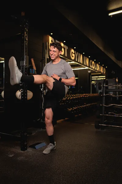 Young man in gray t-shirt laughing while doing a standing high kick stretch inside a dark Colorado gym