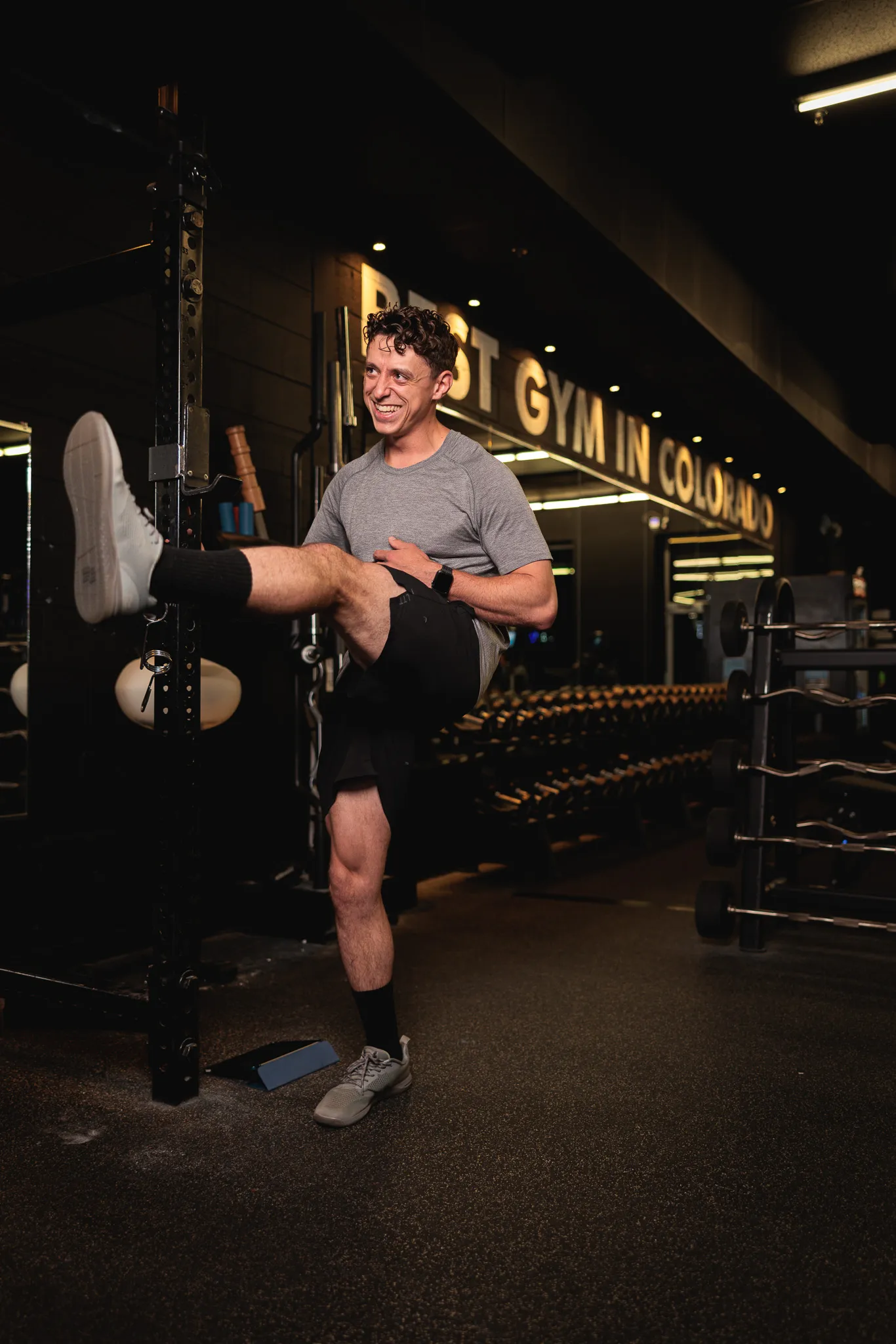 Young man in gray t-shirt laughing while doing a standing high kick stretch inside a dark Colorado gym