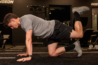 Male fitness trainer performing a bird-dog exercise on hands and knees on a dark gym floor