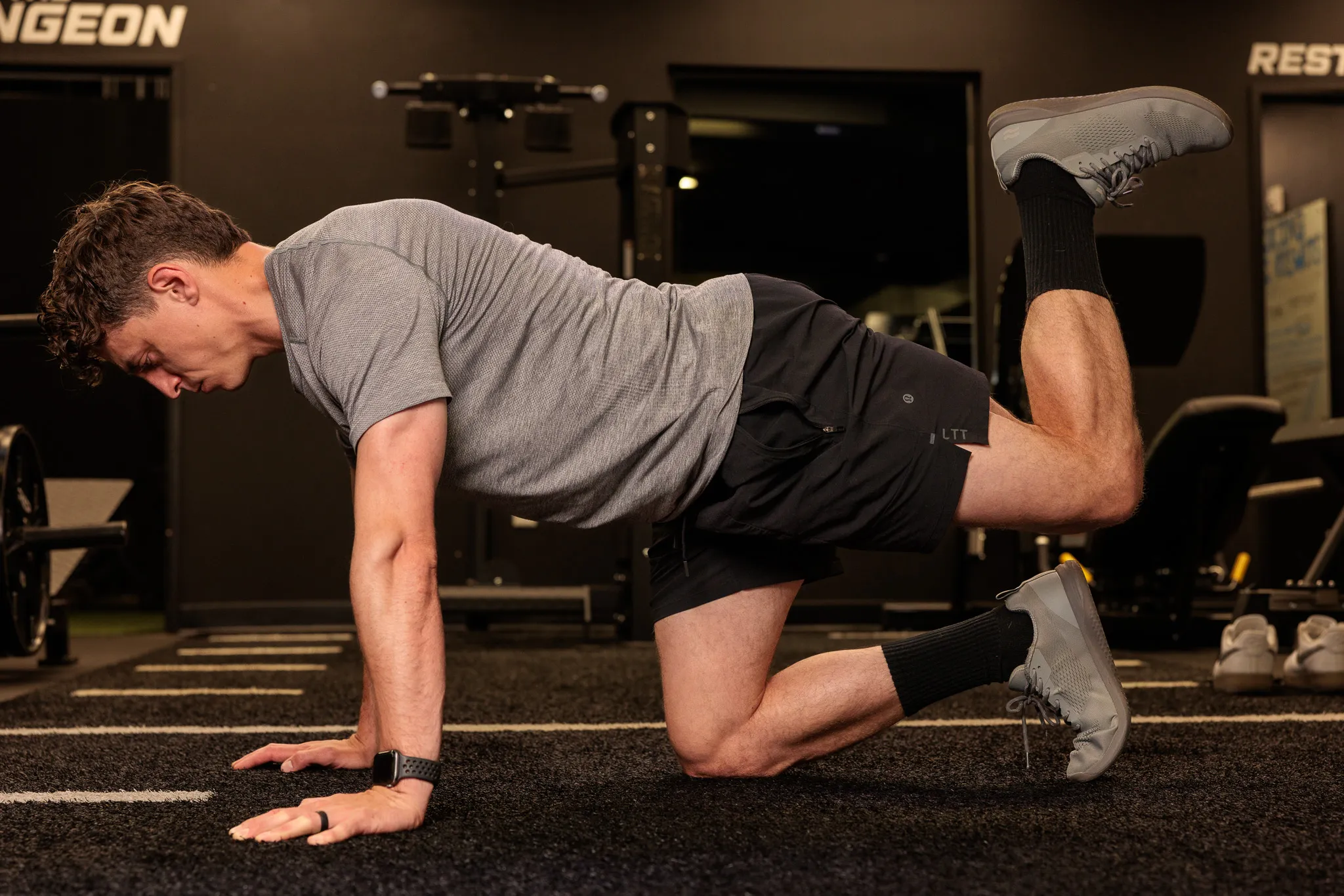 Male fitness trainer performing a bird-dog exercise on hands and knees on a dark gym floor