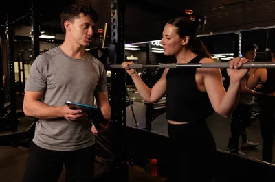 Personal trainer holding a tablet while coaching a woman doing barbell back squats in a dark gym