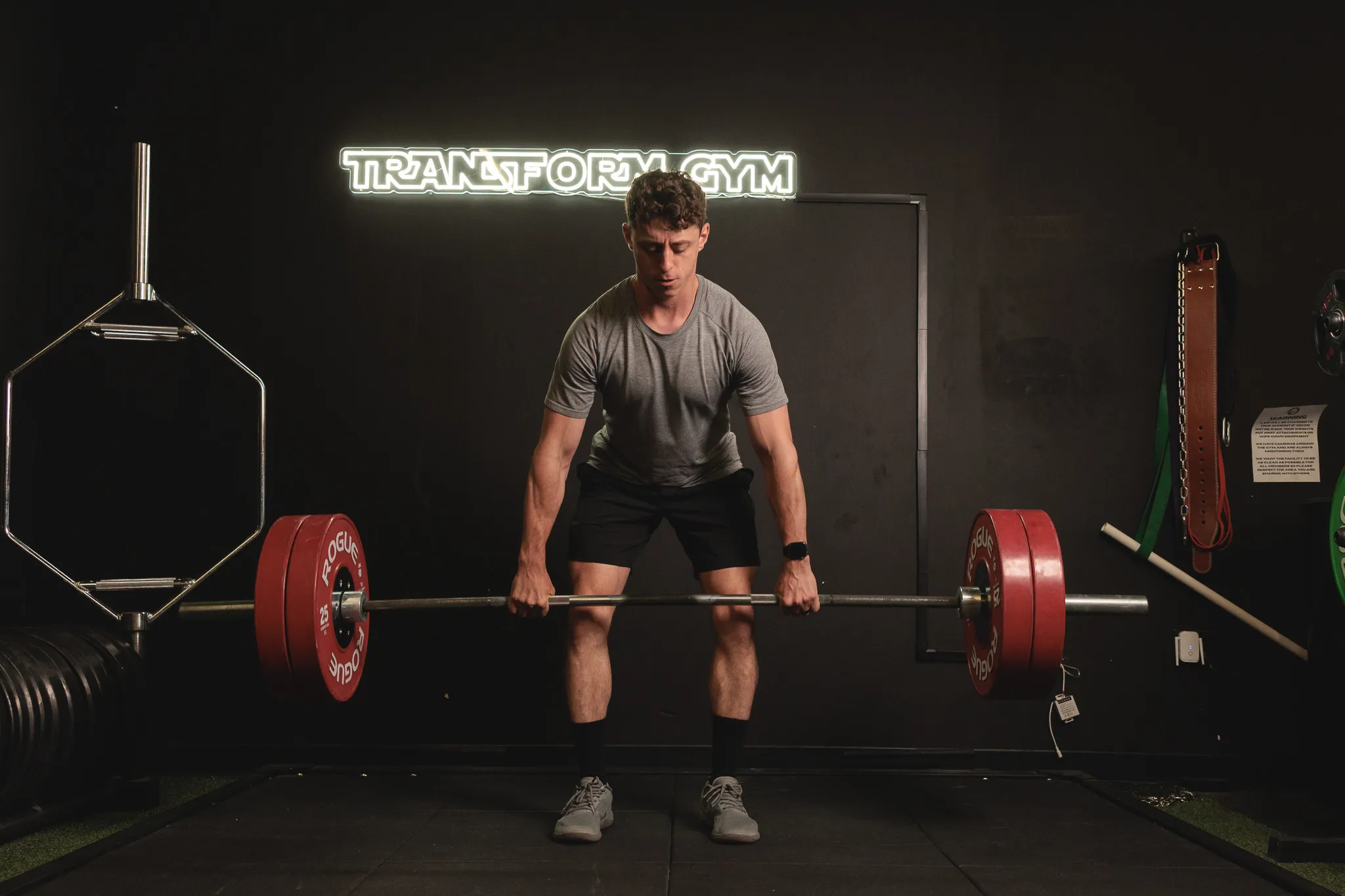 Fitness trainer deadlifting a loaded barbell with red plates under a neon Transform Gym sign