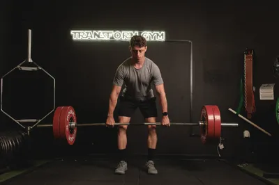 Fitness trainer deadlifting a loaded barbell with red plates under a neon Transform Gym sign