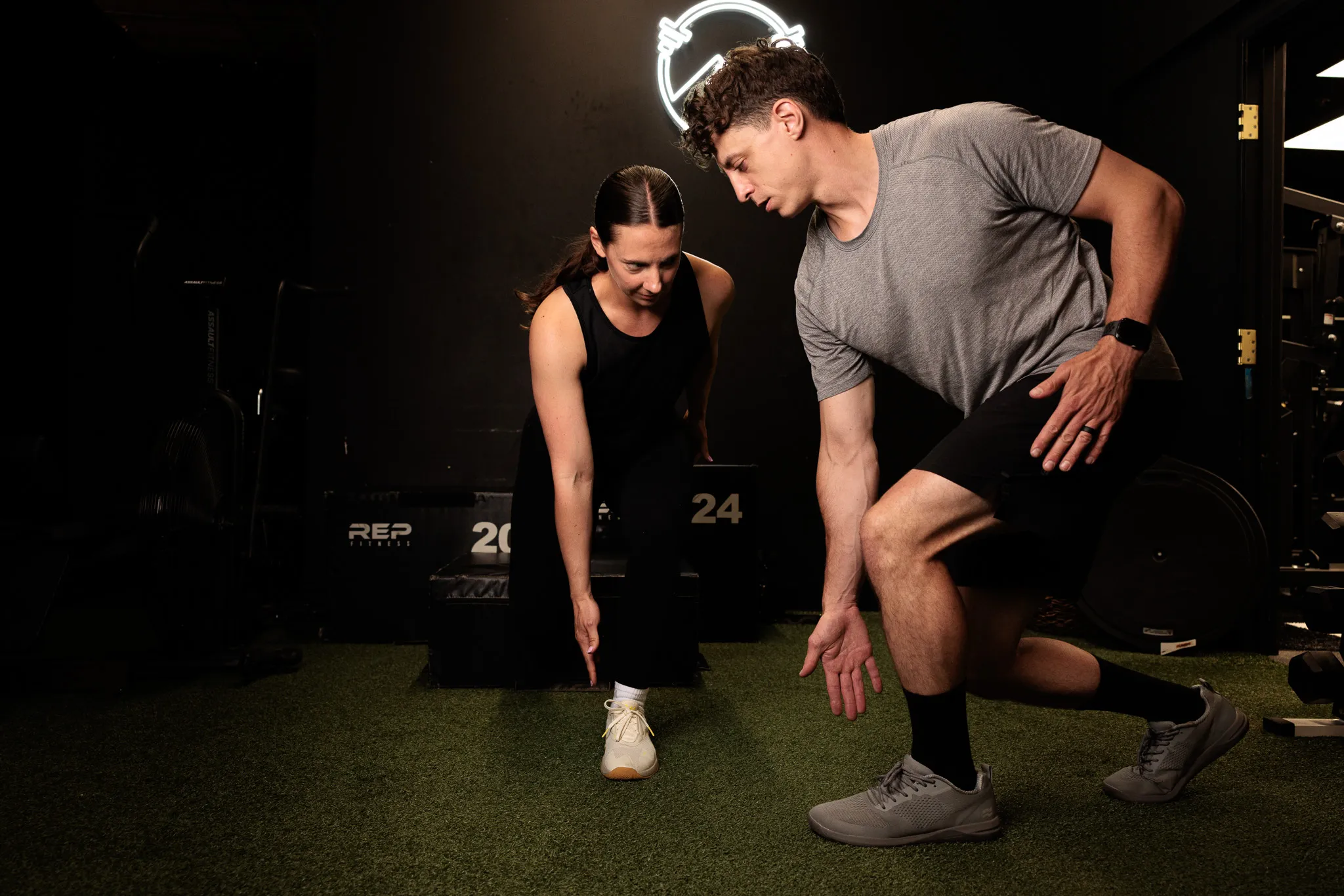 Two fitness professionals in a gym, woman lunging forward while male trainer demonstrates form on green turf
