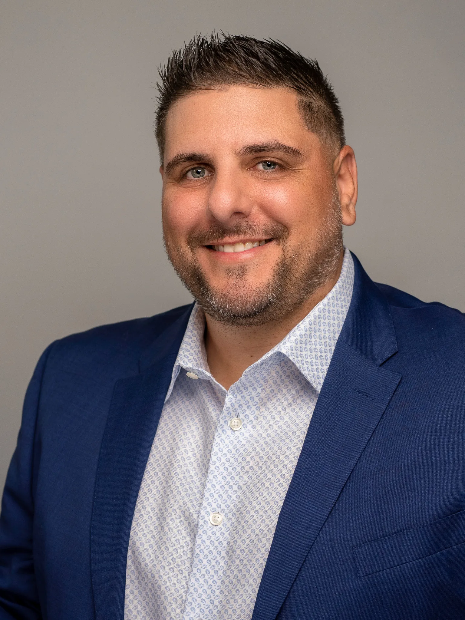 Man in his 30s smiling in navy blue blazer and patterned white shirt against gray studio backdrop, headshot crop