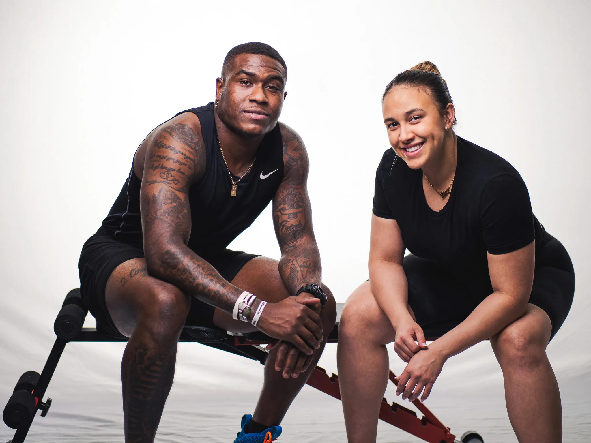 Two young athletes sitting on a weight bench in studio, man with tattoos and woman smiling in black workout gear
