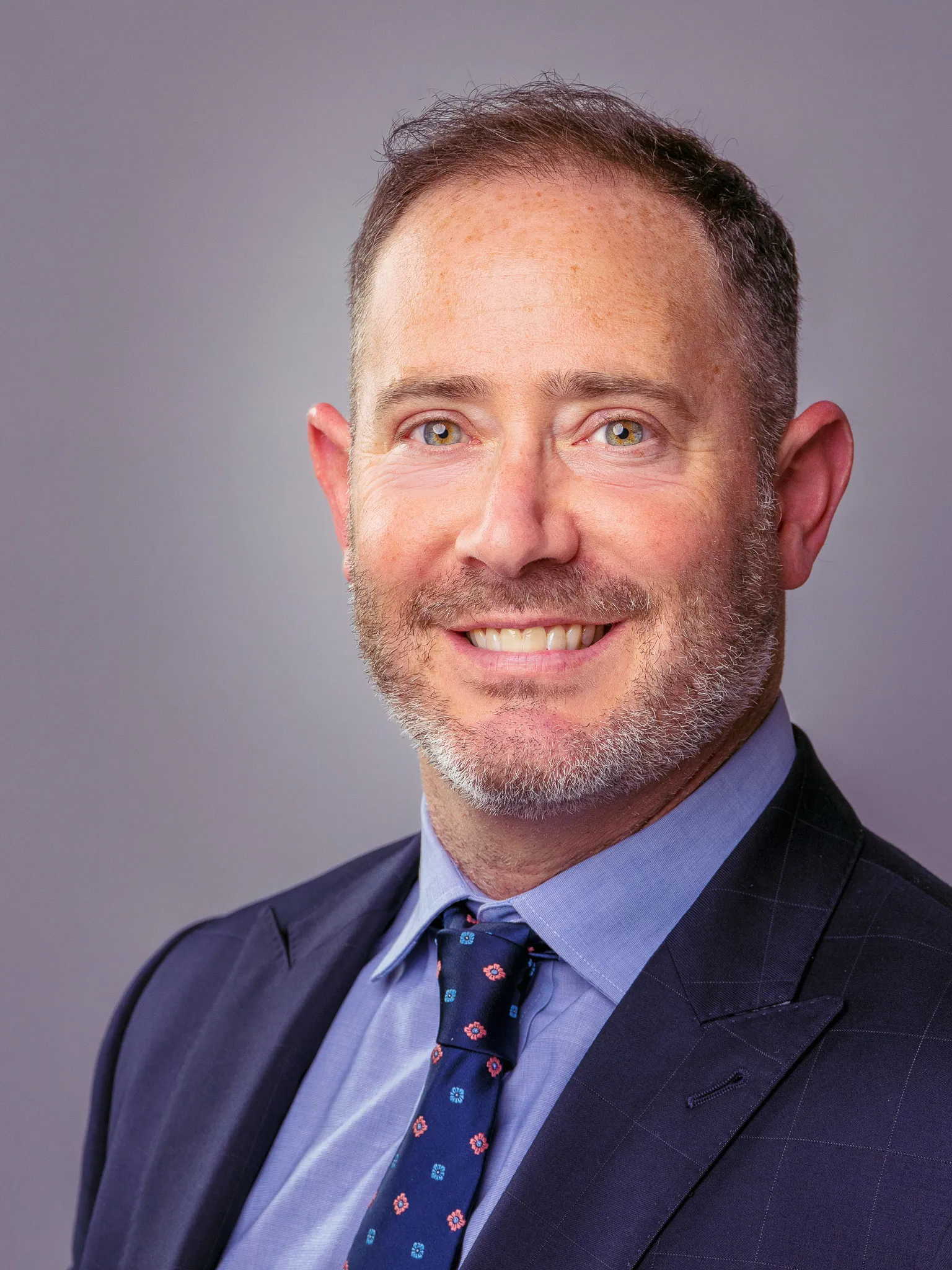 Middle-aged man smiling in navy suit and patterned tie against gray studio backdrop, professional headshot
