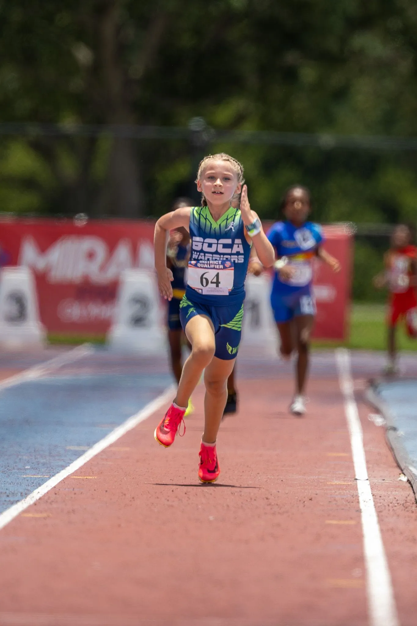 Young girl in Boca Warriors jersey sprinting down the track at a district qualifier meet