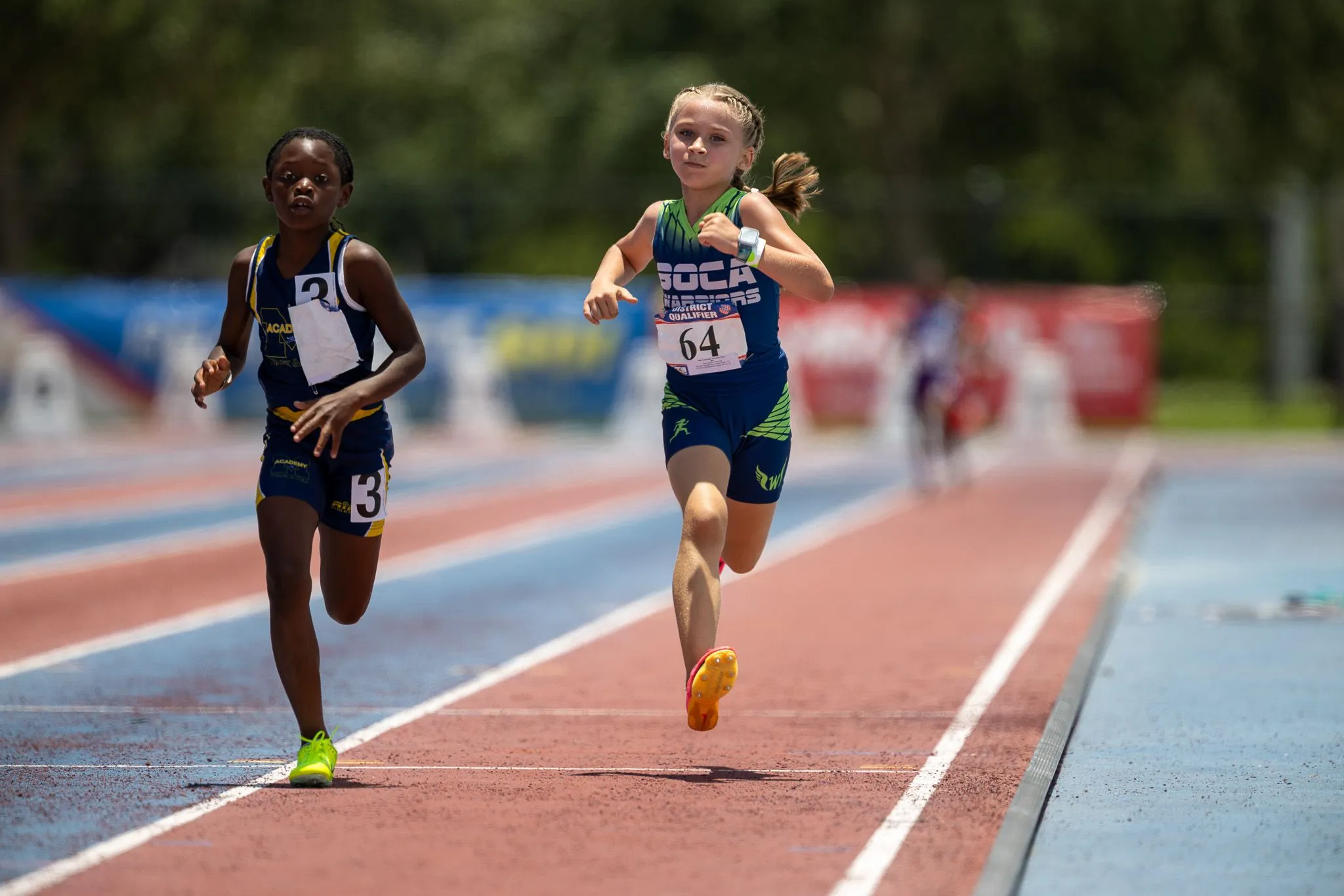 Two young runners racing side by side on a track at a youth district qualifier event