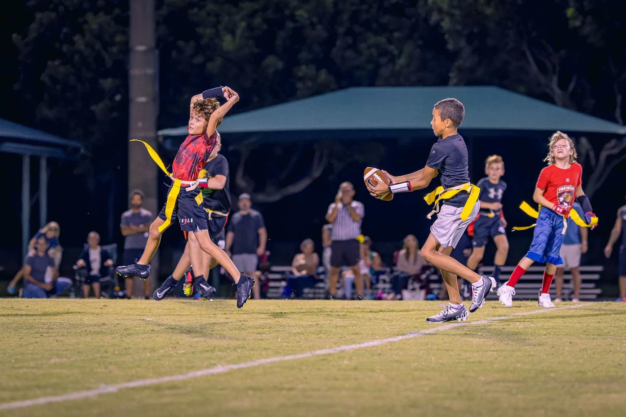 Youth flag football player catches the ball while another leaps to pull a flag at night