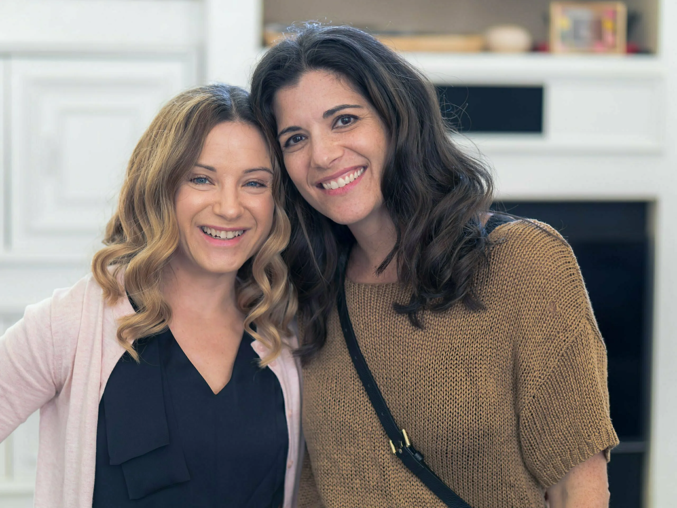 Two women smiling together in a bright kitchen, one in pink cardigan, one in tan sweater
