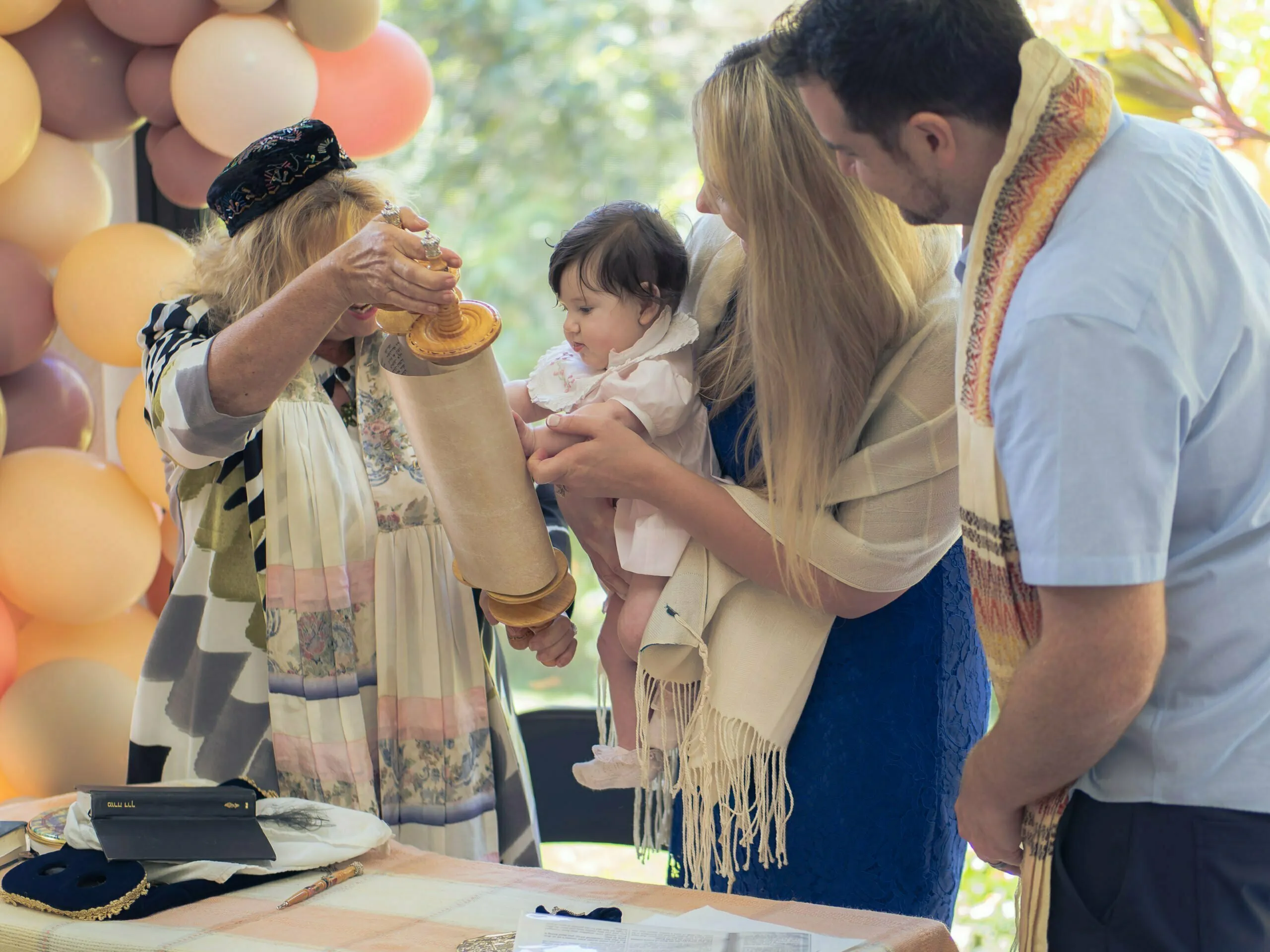 Rabbi performing baby naming ceremony outdoors with parents holding infant near balloon arch and Torah scroll