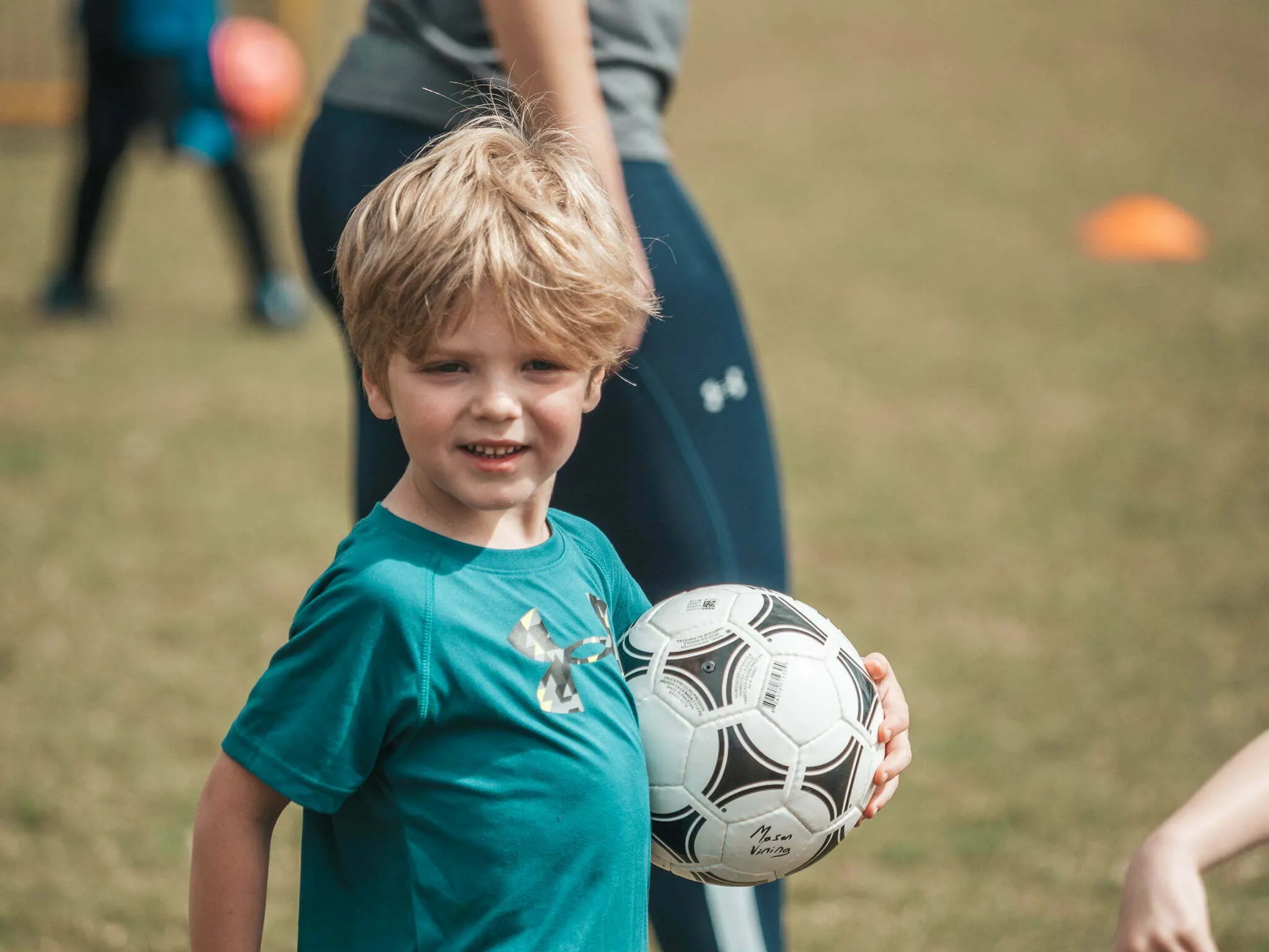 Young blonde boy in teal jersey smiling and holding a soccer ball on a grass practice field