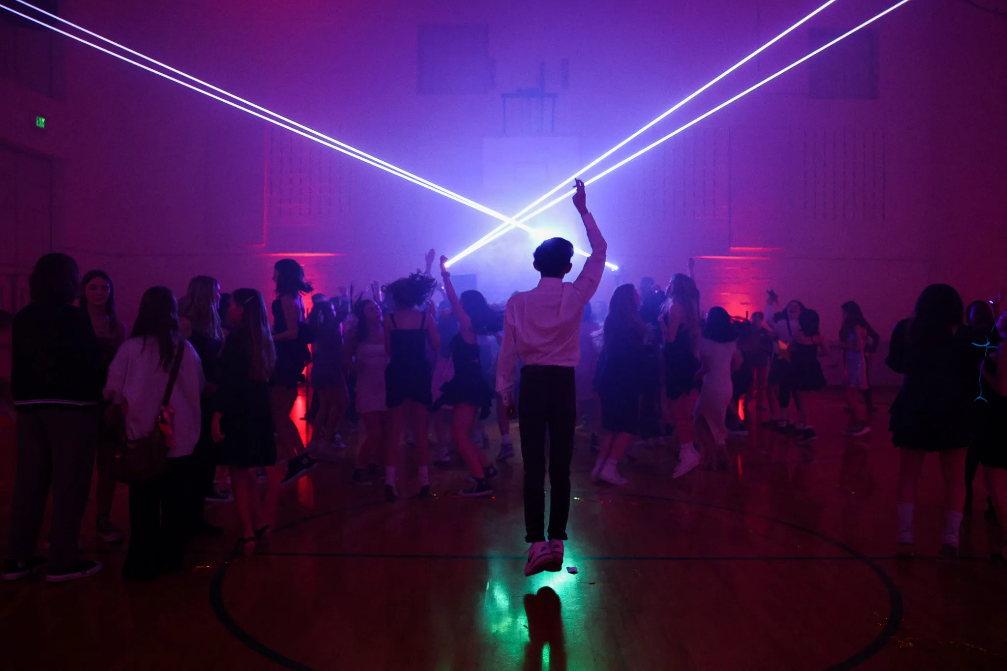 Teen standing center of gymnasium dance floor with arms raised, purple laser lights, and crowd of students