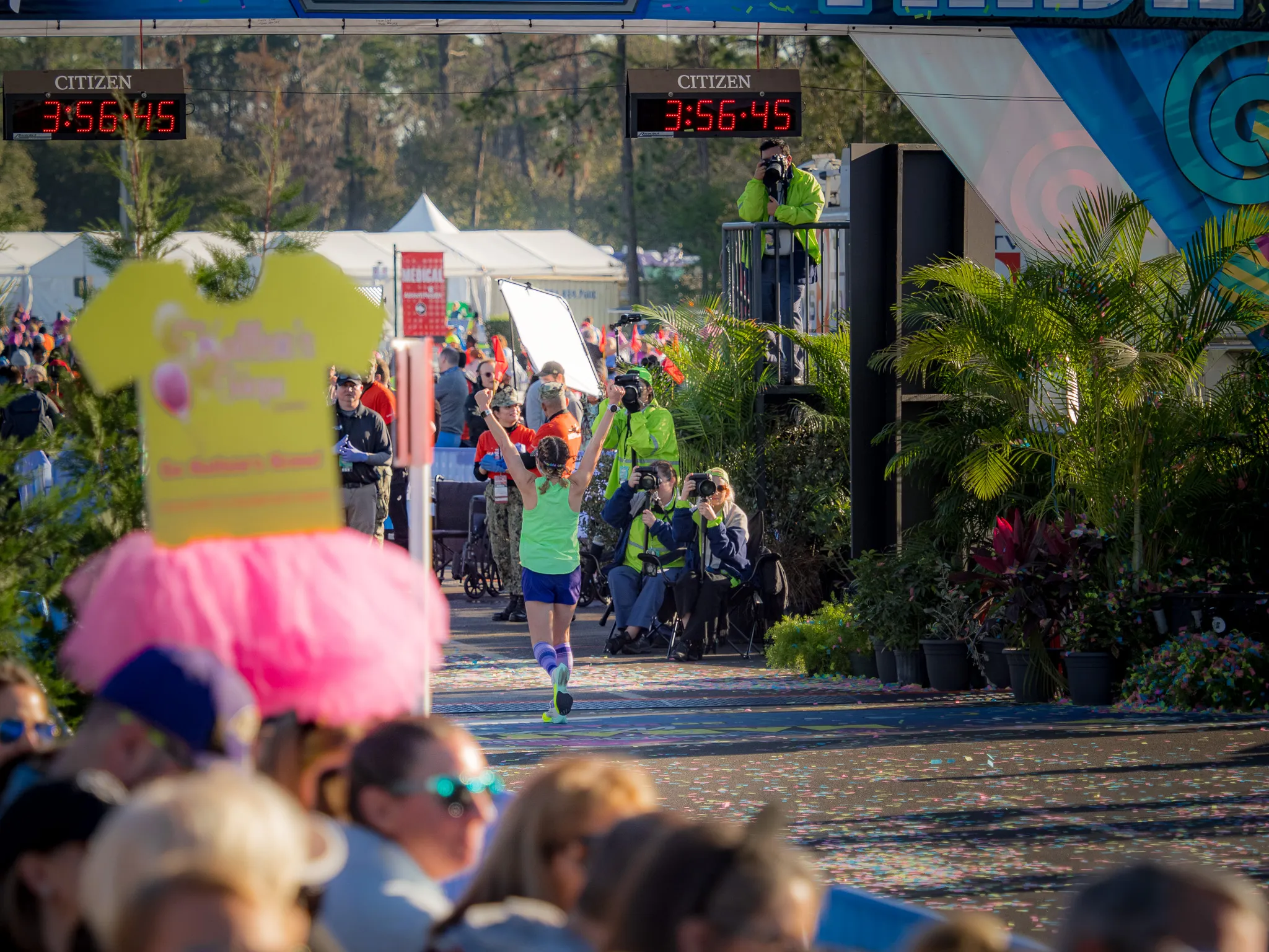 Runner in green shirt crossing marathon finish line at 3:56:45 with cheering crowd and Citizen clocks overhead