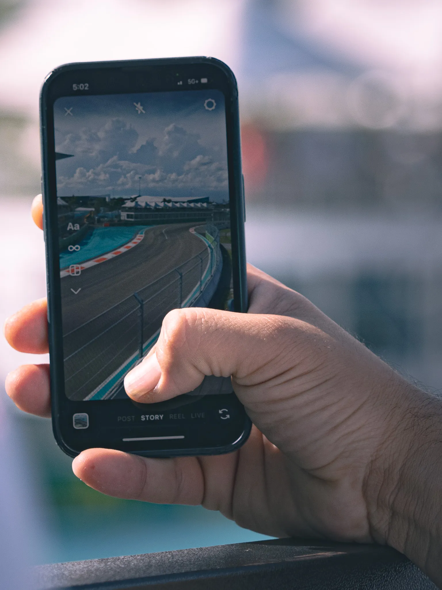 Hand holding a smartphone displaying an Instagram story of a Formula One race track with blurred grandstands behind