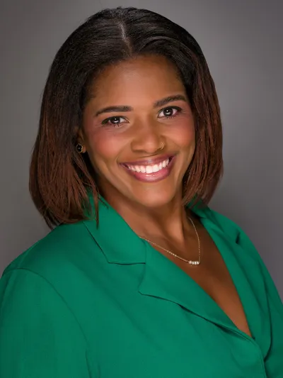 Woman in a green blazer smiling warmly for a professional headshot on a gray background