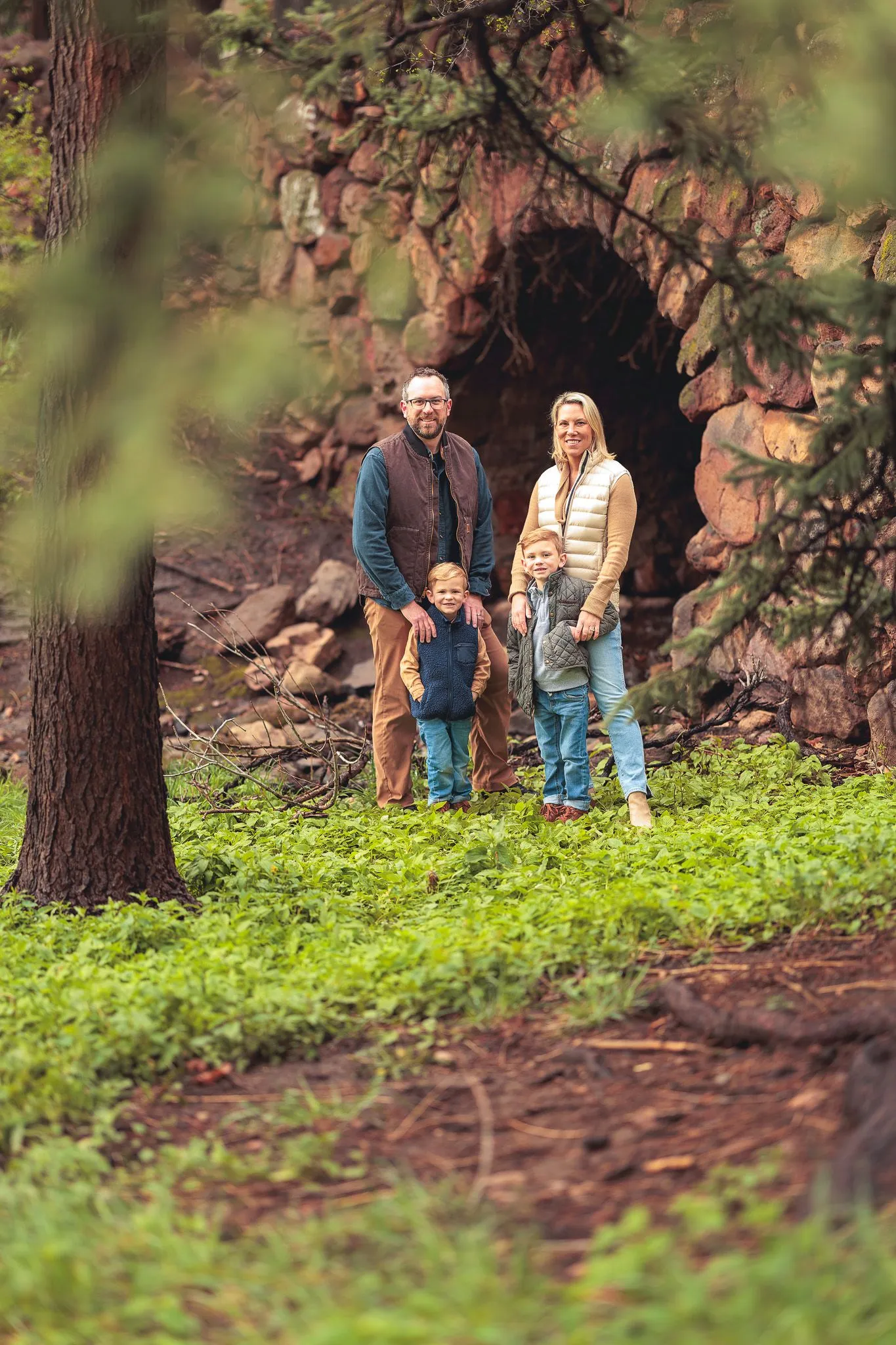 Family of four framed by pine trees standing near a red rock cave in a forest clearing